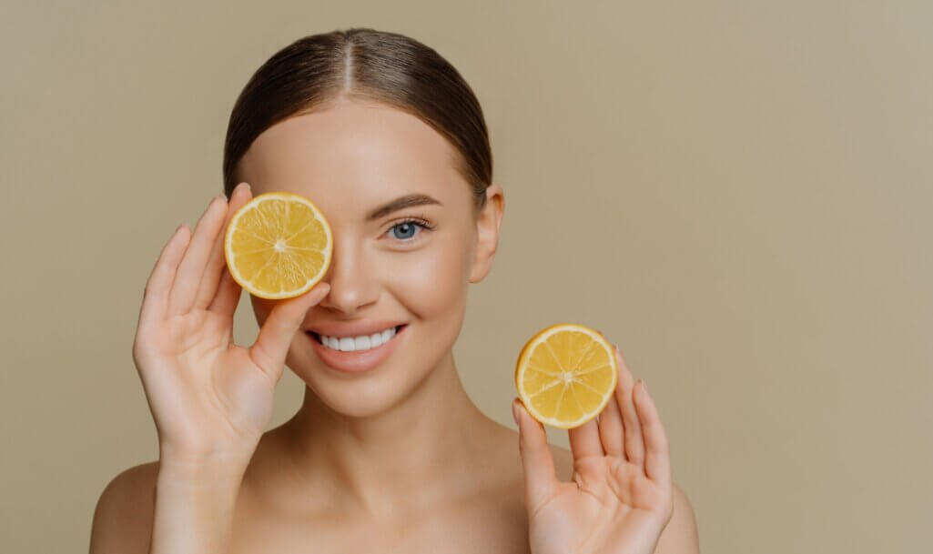 Isolated shot of good looking tender brunette woman smiles gently at camera covers eye with half of lemon holds favorite fruit poses topless indoor against beige background.
