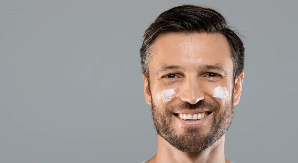 Morning routine. Young happy man with face cream on his cheeks, grey studio background, copy space. Handsome man with face cream or lotion smiling at camera, beauty products for men, male cosmetology