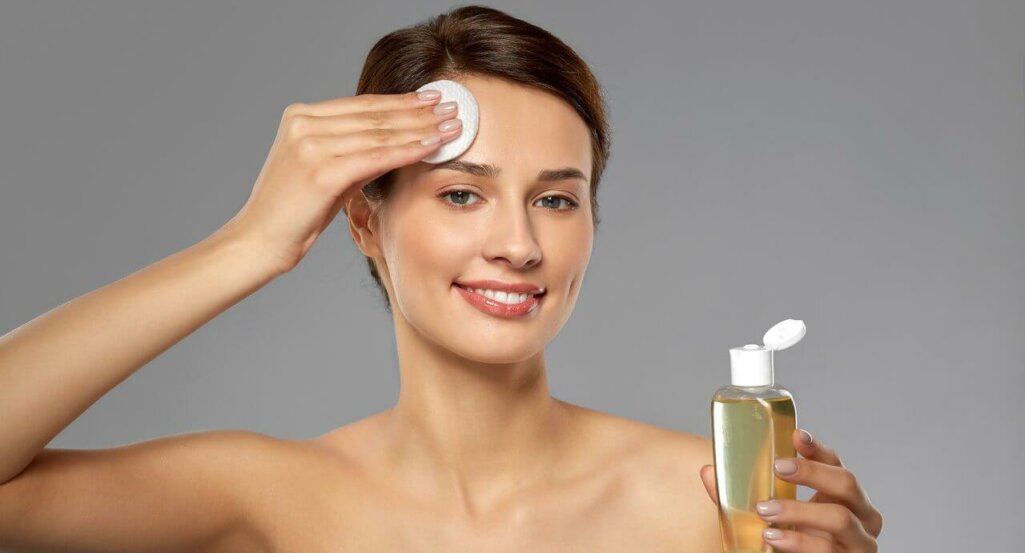 smiling young woman with toner or cleanser and cotton pad cleansing face over grey background