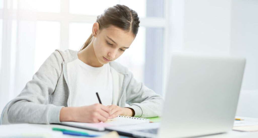 Concentrated teenage girl making notes in her notepad, using laptop