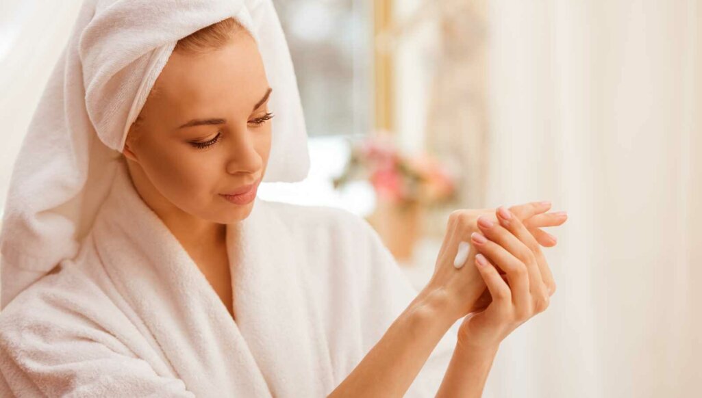Beautiful young woman in a bathrobe with a towel on her head applying cream on her hands, standing in the bathroom