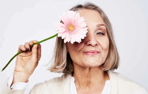 Χαρούμενη κομψή ηλικιωμένη γυναίκα που κρατά ένα λουλούδι κοντά στο πρόσωπο σε γκρίζο φόντο
