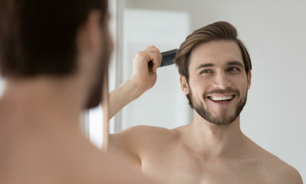 Happy handsome young shirtless man combing smooth straight hair, looking in mirror