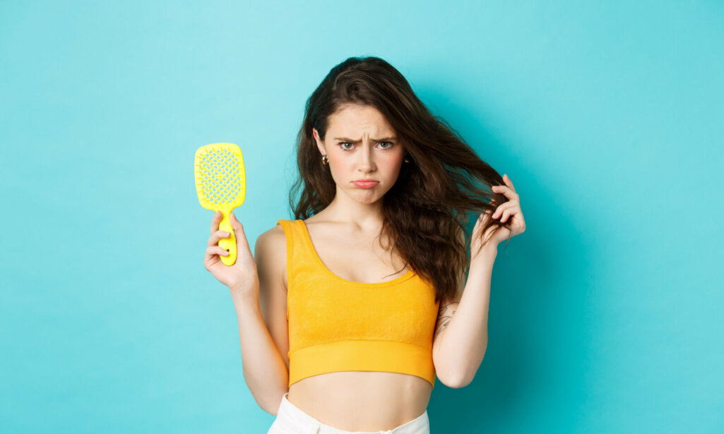 Sad young woman showing brush with strands, having hair loss problem, need cosmetic beauty treatment, standing over blue background upset