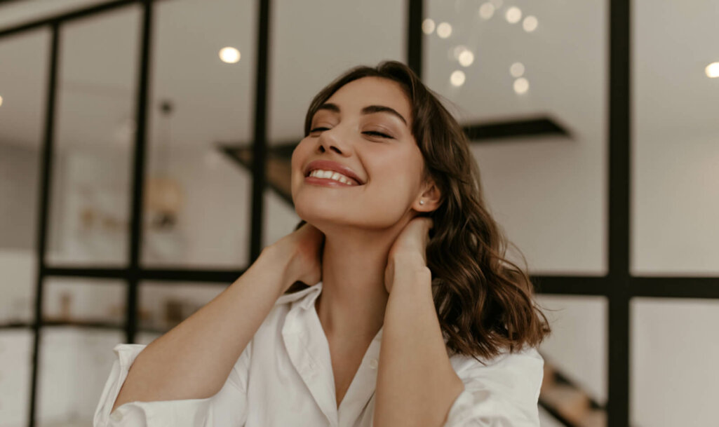 Calm happy brunette woman in cotton white shirt touches neck and smiles. Close up portrait of curly lady in light blouse poses at home. Treatment for migraine