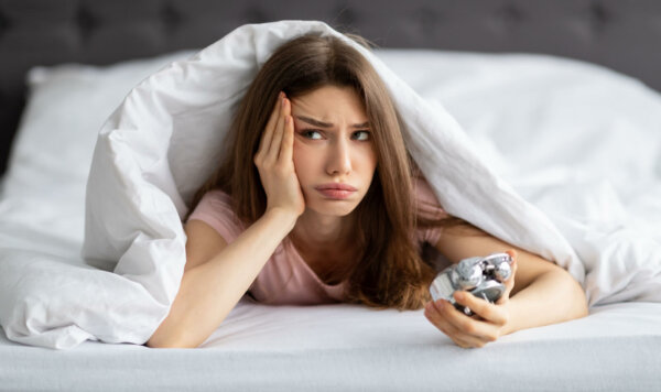 Unhappy young Caucasian woman lying in bed with alarm clock, not willing to get up in morning.