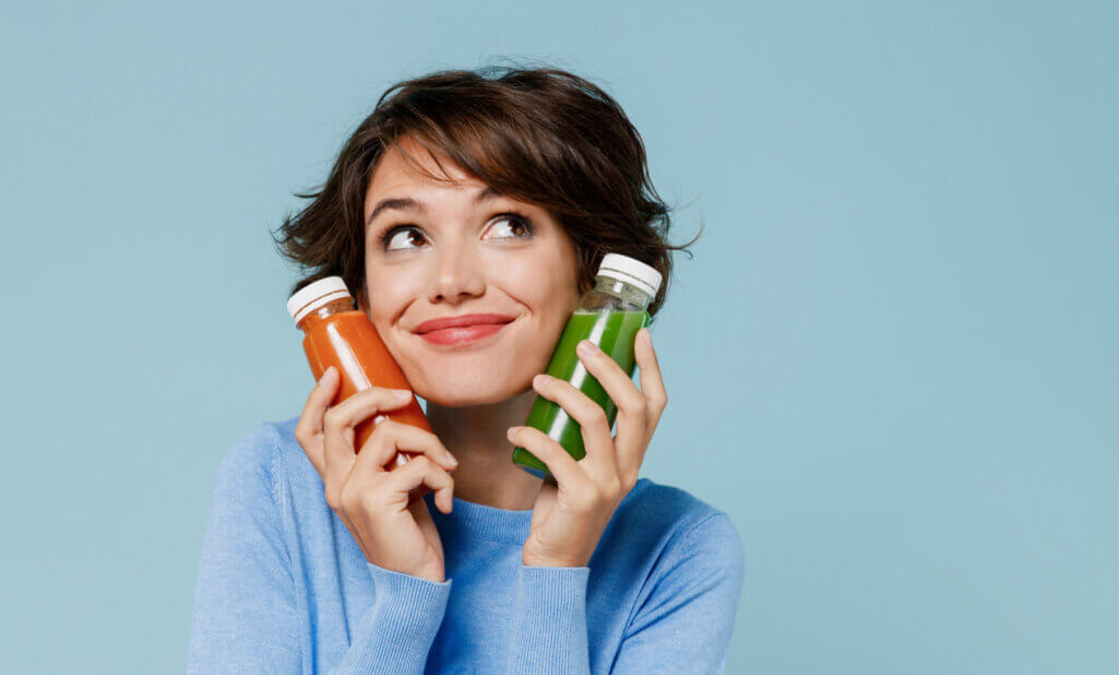 Young dreamful woman in casual sweater hold pressed juice green orange vegetable smoothie as detox diet