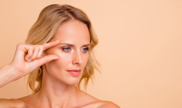 Κοντινό πορτρέτο γοητευτικής γυναίκας με λάμψη και φρέσκο δέρμα δείχνει τα χέρια της κοντά στα μάτια μετά από χρήση κρέμας ματιών για ρυτίδες στα μάτια