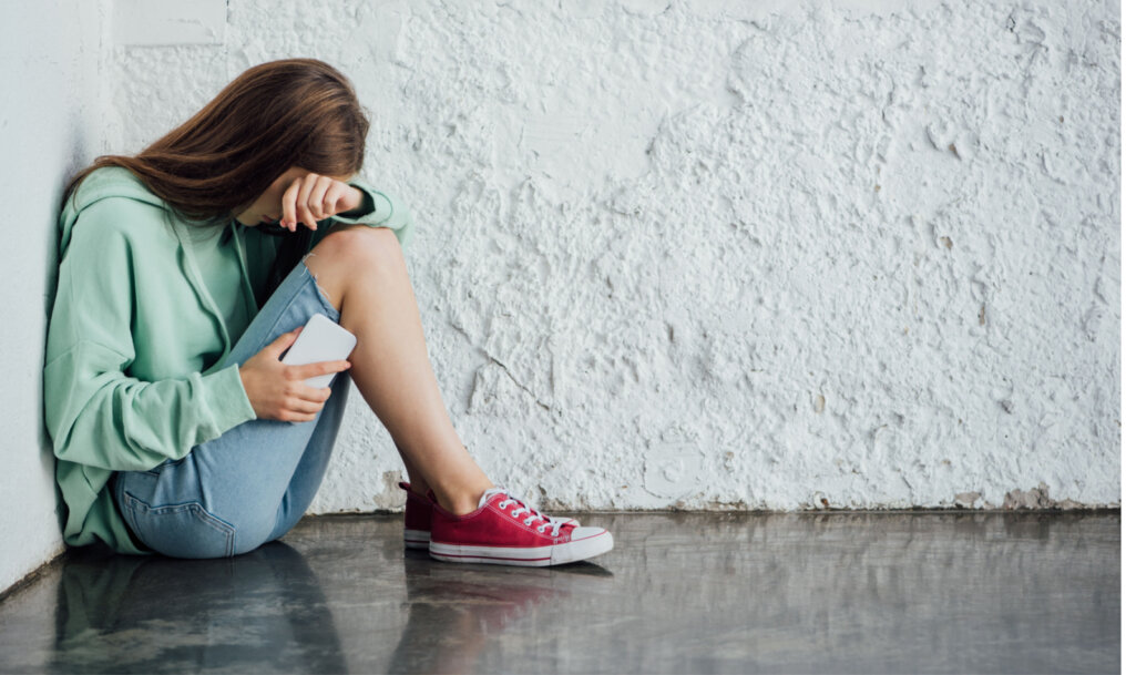 sad crying girl sitting near textured wall and holding smartphone. Cyber bully consept