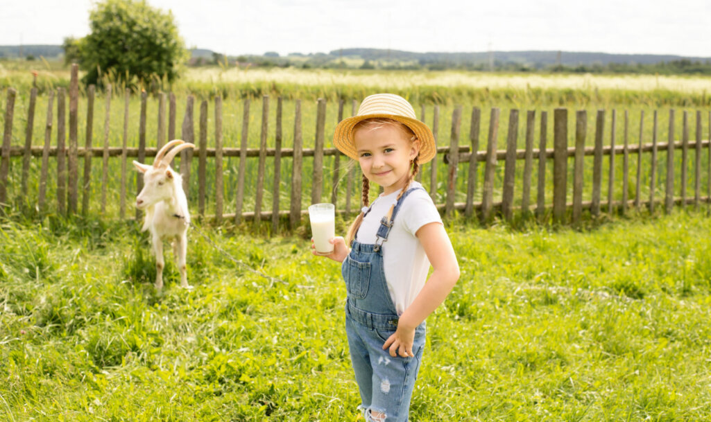 Μικρό κορίτσι κρατάει ένα ποτήρι με κατσικίσιο γάλα για να πιεί και από πίσω της βρίσκεται μία κατσίκα