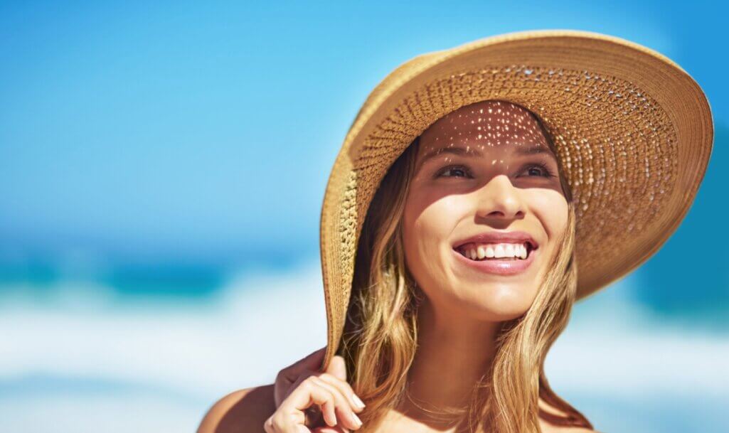 Νεαρή ξανθιά γυναίκα φοράει καπέλο και αντηλιακό προσώπου και χαμογελάει ενώ βρίσκεται στη θάλασσα.