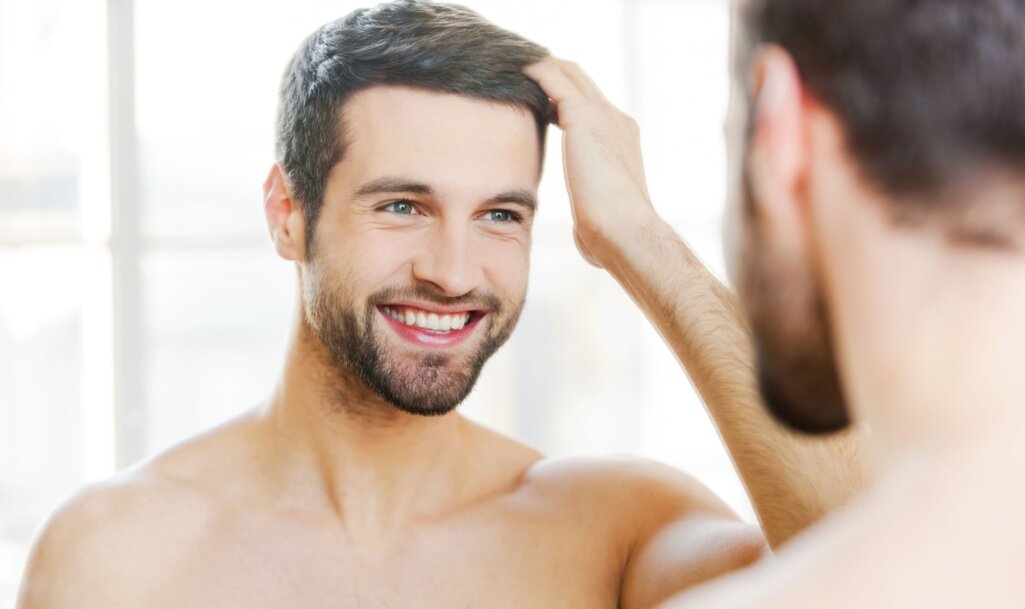 Νεαρός άνδρας χαμογελάει στον καθρέπτη παρατηρώντας τα μαλλιά του αφότου τα έλουσε με ειδικό σαμπουάν